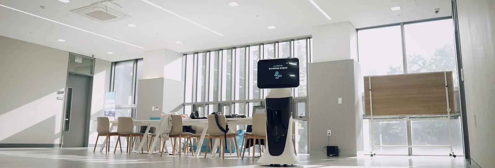 A photo of the patrol robot standing in a large common area of a facility. The robot has a screen on top, with tables and chairs around it.